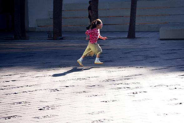 Child with braided hair wearing a pink shirt and light pants running across a sunlit tiled surface. Shadows of trees and other elements fall across the ground. A man stands in the background near concrete steps.