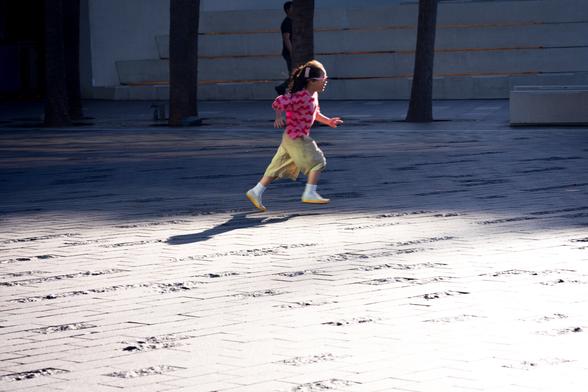 Child with braided hair wearing a pink shirt and light pants running across a sunlit tiled surface. Shadows of trees and other elements fall across the ground. A man stands in the background near concrete steps.