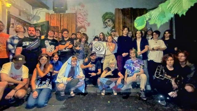 Fotografia grupowa przed budynkiem OCSK Postój. Zgrupowani ludzie trzymają w środku plakat Muzyka Przeciwko Rasizmowi, stowarzyszenia Nigdy Więcej.