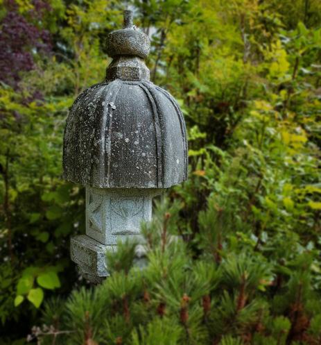 A weathered stone Japanese garden lantern stands amongst vibrant green foliage. The lantern has a bulbous top with a domed cap, a rectangular base with subtle carvings, and is covered in moss or lichen, suggesting age and exposure to the elements. The blurred background features dense foliage in various shades of green, creating a tranquil and natural setting.