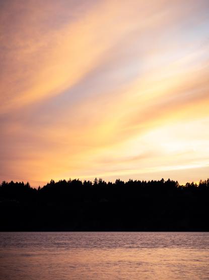 the sunset near Tacoma Narrows Bridge. The sky is orange, purple, and light blue. The land featured in the image is nearly pure black. The water reflects the colors of the sky.