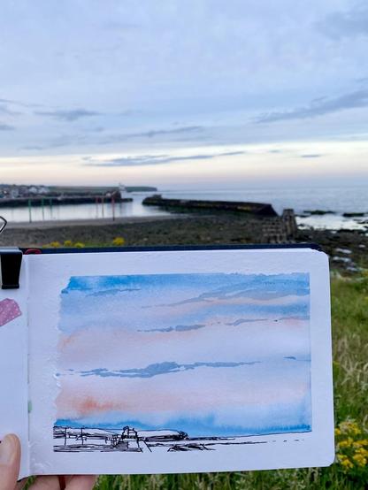 A pale sky over a harbour, in front a sketchbook with a coloured sketch of that scene, but the margins are uneven and ripped.