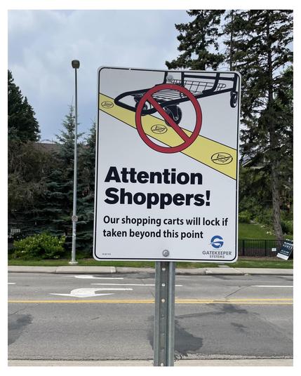 Vertical road sign with a message for shoppers stating "Attention Shoppers! Our shopping carts will lock if taken beyond this point," featured against a cloudy sky. The sign includes a symbolic image of a shopping cart crossed out in red, positioned at the upper half, on a yellow detailed base with black borders. Green trees and a road with a white painted arrow are visible in the background, along with a street lamp to the left. An emblem saying "Gatekeeper Systems" is positioned in the bottom right corner of the sign.