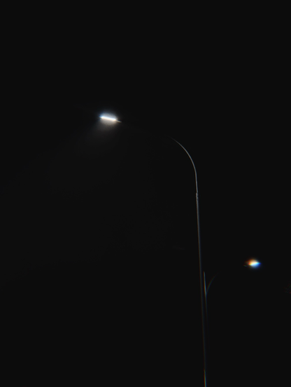 A single streetlamp shines brightly against a black sky, accompanied by another distant lamp creating a subtle colored glow