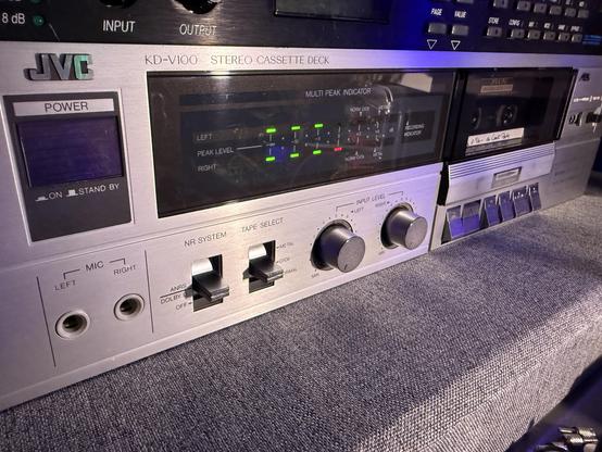 The front of a large cassette deck recorder with playback from the 1980's that is bright chromes and silvers, across the front is a level meter in green and red lights. The blocky metallic-looking logo on the far left reads: JVC