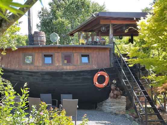 bateau ancien, probablement chinois, posé sur le sol derrière 4 chaises de jardin. Des escaliers en métal permettent de monter sur le pont. On voit 4 fenêtres carrées dans la coque en haut, une bouée de sauvetage orange accrochée à l'extérieur, un tonneau est posé à l'avant vers le mât. Il y a de la verdure tout autour, dont des arbres.