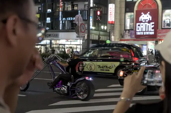 Photograph of a biker riding a purple-and-chrome motorbike with an unusally long front fork and handlebars. The bike rider is wearing a purple retro-looking half helmet, a black T-Shirt with a large Che Guevara graphic on the back and black trousers.·
He is riding his motorbike in the middle of a Shinjuku street with busy building lights around. The photograph is taken from between two people watching the scene, their faces blurry because of their proximity and the focal length. The person on the right is holding her camera horizontally to capture the scene.