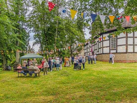 The photo depicts a social gathering or community event taking place outdoors during daylight hours. Several people are standing around chatting, some seated at tables with food and drinks under a green canopy tent, while others walk through the area. The setting includes traditional buildings with timber framing, hinting at historical significance or cultural heritage. There is festive decoration in the form of colorful triangular flags strung across trees above, adding to the celebratory atmosphere. Bicycles are parked nearby on the grass, suggesting attendees have cycled to this location. Overall, it appears to be a relaxed and friendly event with an emphasis on community engagement.