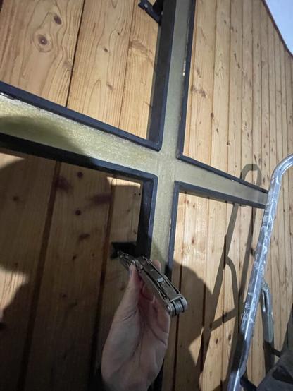 The image shows a close-up view of someone's hand holding an Leatherman multitool against a wooden wall with a metal cross. The wood has a natural grain pattern typical of pine or similar softwoods. There are four prominent cross-shaped cut-outs, likely for hinges or locks, and one is being interacted with by the person in the photo. The individual appears to be using what looks like a multi-tool or pliers on this particular metal reinforcement structure of the cross. The lighting casts shadows that create an interesting pattern against the wood grain. On the right side of the image, part of a metallic ladder is visible.