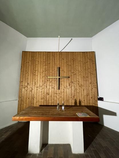 This image shows a room with minimalistic decor focused around religious symbolism. The central feature is a large wooden wall adorned with a cross made of metal or another reflective material. Above this wall hangs an elongated object that appears to be a rod, possibly part of lighting equipment. In front of the wooden wall stands a simple rectangular table on two white supports. The room has plain walls painted in light colors with one corner showing a darker greenish hue on the ceiling. There is also an electrical outlet visible to the right side of the image. The overall setting suggests this could be part of a chapel or prayer space designed for contemplation and reflection, given its clean lines, lack of decoration aside from essential religious symbols, and serene atmosphere.