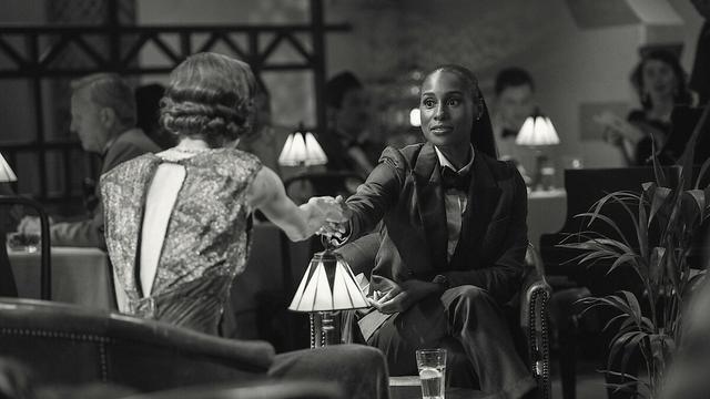 Schwarz-weiß-Szene aus Black Mirror – Hotel Reverie: Zwei Frauen geben sich in stilvoller Bar-Atmosphäre die Hand. Links sitzt eine Frau mit elegantem, rückenfreiem Kleid, rechts eine Frau im Anzug mit Fliege. Die Beleuchtung ist sanft, die Stimmung wirkt nostalgisch und träumerisch.
Foto: Netflix