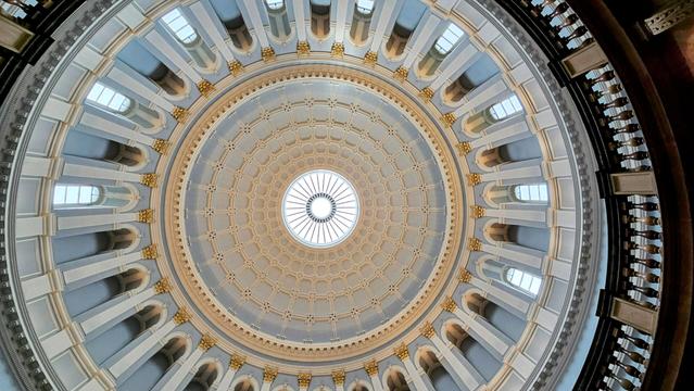A low-angle, wide-shot photograph captures the interior of a majestic, circular domed ceiling. The dome is predominantly light grey and white, with intricate gold detailing along the decorative arches and ledges. Multiple rectangular windows are evenly spaced around the base of the dome, allowing natural light to filter in. At the very centre of the dome, a circular skylight with a radiating spoke pattern is visible. To the right of the frame, a curved, dark banister or railing is partially visible, hinting at an upper level or staircase within the building. The overall impression is one of architectural grandeur and symmetrical beauty.