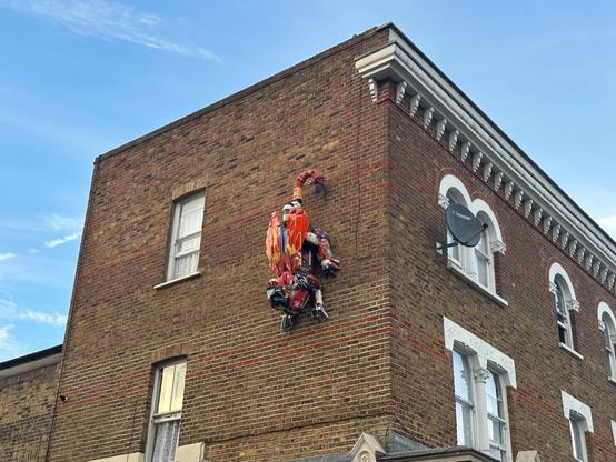 The side of an old London apartment block, with a large red insect/scorpion-shaped sculpture made of reclaimed metal/plastic parts