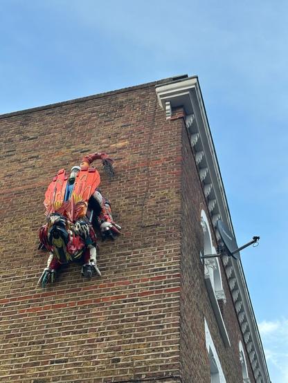 a large red insect/scorpion-shaped sculpture made of reclaimed metal/plastic parts, on the side of a brown brick building