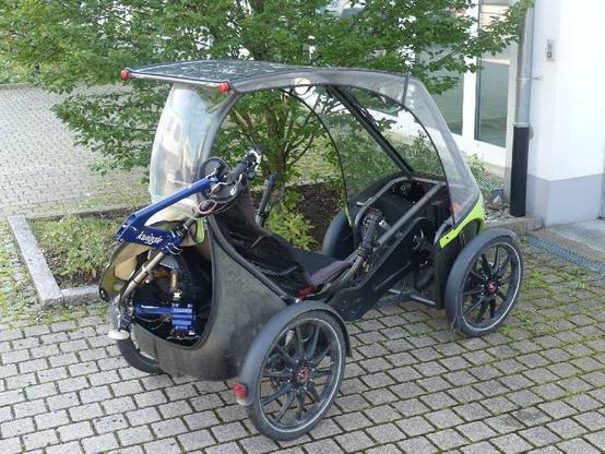 Ein Pedilio (Solarvelomobil) mit einem halb gefalteten Kwiggle (Faltrad) im Kofferraum, das Kwiggle ragt etwas heraus, vor einer Einfahrt stehend