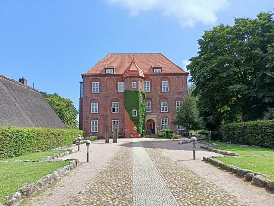We see the front of Agathenburg Castle, a huge brick facade and a road that points towards the entrance. The picture was taken around midday, the sun shines bright and the sky is blue.