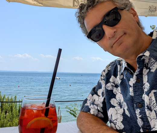 Sul lato destro della foto un uomo in primo piano con gli occhiali da sole e una camicia bianca e blu floreale sul lato sinistro in calice con dentro una bevanda rossa, spritz con il Campari sullo sfondo il lago di Garda