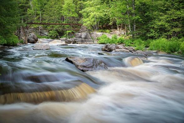 The blurred water of Duchesnay Falls flows down a river, under a stone footbridge through a forest in North Bay, Ontario during sunset.