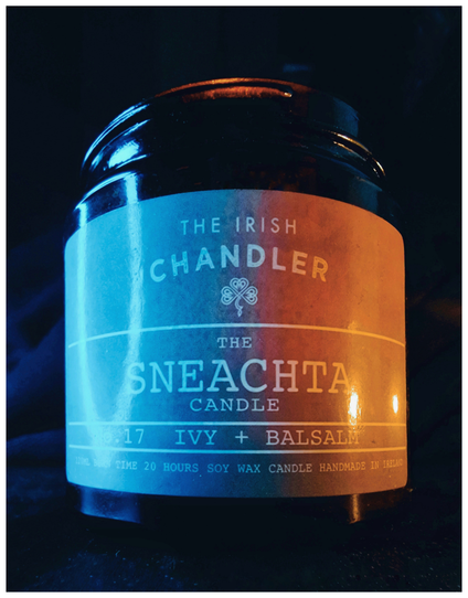 This photo is a close-up of a candle labeled "The Irish Chandler, The Sneachta Candle, No. 17 IVY + Balsam" in a black glass jar. The label also features at the bottom - "120ml burn time 20 hours soy wax candle handmade in Ireland ".The candle has a blue and orange gradient due to different coloured lights which are to the sides of the candle. The background is dark, highlighting the candle's colours and design.