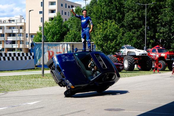 Ein blauer BMW der seitlich auf zwei Rädern fährt. Darauf steht ein Mann der auf dem Auto „surft“ und eine Gruß Geste macht. 

Im Hintergrund Gebäude, Bäume, ein weißer und ein roter Monstertruck