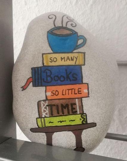 Bemalter Stein mit einem fünf Büchern auf einem braunen Tischchen.
Die Bücher sind gelb, blau, orange, braun und hellgrün.
Verteilt auf die Bücher steht der Spruch:  'So many books so little time'.
Auf dem obersten Buch steht eine blaue Kaffeetasse mit Dampfwölkchen.