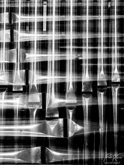 Photographie monochrome en format vertical d'une double exposition des tuyaux d'un orgue : première captation du bas des tuyaux en format horizontal et seconde exposition en basculant le boîtier à 90°. Les tuyaux se croisent à 90°, de même que les vagues des ouvertures générant le son (comme sur un sifflet), plus ou moins éloignées de l'extrémité des tuyaux selon leur diamètre.