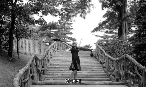 Black and white image from the 1962 film Cléo from 5 to 7, directed by Agnès Varda. It shows Corinne Marchand as the singer Cléo, pausing on her way down a series of wide wooden steps in a park. She has thrown her arms dramatically to the sides, extending a scarf, her hips slightly twisted, as she says, "Music!" She has blonde hair and is wearing a dark, knee-length dress. Tall trees grow on either side of the steps.