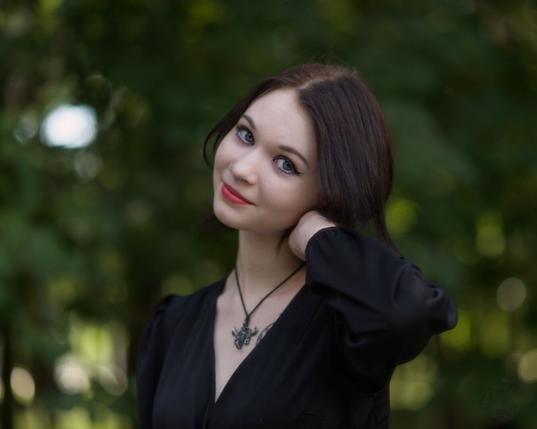 The image captures a young woman with long brown hair in an outdoor setting, surrounded by greenery. She is wearing a black dress and a necklace. Her red lipstick stands out against her attire's dark hue. The background suggests she might be near trees or bushes with leaves visible around the edges of the frame. Her posture exudes confidence; her left hand rests on her neck while the other hangs down, creating a relaxed yet poised stance. She is gazing directly at the camera. The lighting in the photo appears natural and soft, hinting at an outdoor shoot during daylight hours with no harsh shadows visible on her face or body.

In terms of style, she has opted for minimal jewelry, choosing instead to wear what seems like a pendant necklace. Her hair is styled straight down past her shoulders, which complements the formal look but also adds a touch of casual elegance due to its lack of intricate styling.
