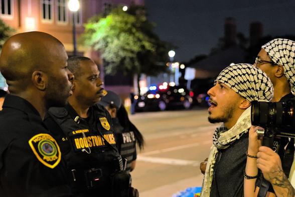 Three activists facing off against the police outside of the Harris County Jail at night, illuminated only by street lights, chanting in support of an unjustly jailed comrade being detained inside. The police are stone-faced and ignoring the activists' eyes staring directly at them.