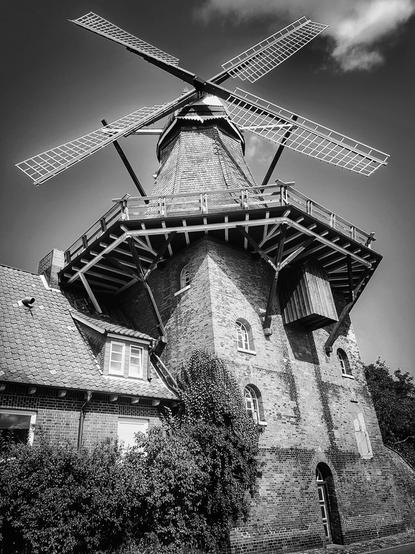 A dramatic, low-angle black and white shot of the historic Jork windmill in Germany. The brick windmill stands prominently with its large sails reaching towards a partly cloudy sky. Intricate wooden details adorn its upper structure, and a smaller attached brick building is partially visible at its base, surrounded by green foliage.