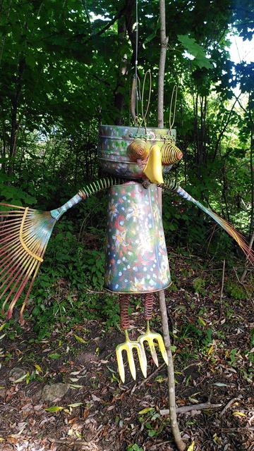 Sculpture métallique colorée représentant une sorte de hibou suspendue au milieu des arbres. Ses ailes sont faites à partir de râteaux, ses pattes à partir de petits outils de jardinage à dents et le reste à partir de matériaux de récupération agencés et peints.