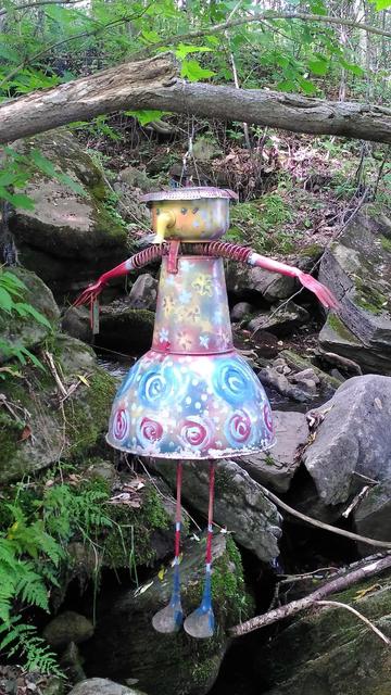 Sculpture en métal peint représentant un personnage suspendu à une branche dans les bois. Sa tête est une théière, ses bras sont des râteaux rouges, les autres parties sont aussi en matériaux recyclés ou détournés