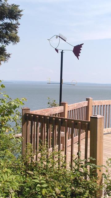 Belvédère en bois qui donne sur le fleuve Saint-Laurent. Une girouette en forme de poisson en métal recyclé surplombe la barrière du belvédère et se découpe sur le ciel bleu.