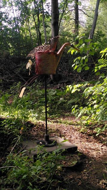 Sculpture en forme d'oiseau rouge aux ailes déployées se détachant sur un fond forestier. Le corps de l'oiseau est fait à partir d'un arrosoir en métal peint et ses ailes à partir de râteaux.