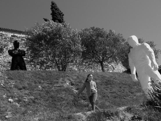 This is a view of a terraced Mediterranean garden with olive and cypress trees, as well as an art installation. Two statues of men in threatening poses are visible in the image: one black on the upper terrace and one white on the lower terrace. A nonchalant little girl walks by on the path. (Version françæ) Vue d’æn jardin méditerranéæn en terrasse avec des oliviers, æn cyprès, mais surtout æn installation artistique. On voit sur l’image deux statues d’hommes massifes aux postures menaçænt, æn noirx sur læ terrasse dal haut et æn blank sur ceil dal bas. Sur læ chemin passe æn petix fille nonchalænt.
