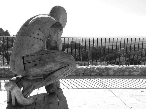 A wooden statue in high heels is seen from behind as e looks down at the plain below. Squatting on the high terrace, e appears ready to bounce. (Version françæ) Statue en bois à talons haux vux de dos, læ regard vers læ plaine en contre-bas. Accroupix, on læ dirait prêx à bondir dal terrasse en hauteur.