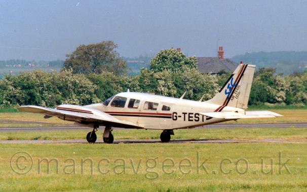 Side view of a twin propellor-engined light aircraft taxiing from right to left along a narrow grey tarmac taxiway.
The plane is a light sandy-brown, with a red and black stripe running along the body and up into the tail, as well as along the engine cowlings mounted on the wings.
The black registration "G-TEST" is overlaid on the stripes on the rear fuselage, with large blue text "SFC" and black text "Air taxis LTD" over the stripe on the tail.
Green grass fills the foreground, with a line of trees in the background, in front of a house with a grey roof.
Grey skies over trees in the distance fill the rest of the frame.
