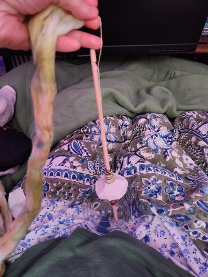 A view of my lap. There's my Indian print bedspread, and a small glass dish in my lap. I'm holding a foot-long wooden dowl that has a funky homemade whorl on it to make a spindle. I've got a small amount of spun cotton already wound onto the dowel, and a length of combed and dyed cotton fiber ready to be spun.