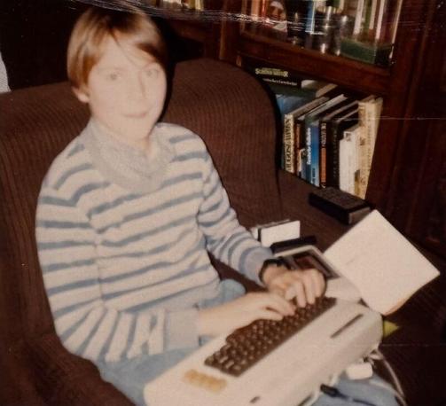 Ein etwa 12-jähriger Junge sitzt konzentriert vor einem Commodore VC20 Heimcomputer. Er trägt ein T-Shirt und hat kurze dunkle Haare. Auf seinem Schoß liegt ein VC20 - ein Brotkasten-förmiger früher Heimcomputer. Die Umgebung ist das elterliche Wohnzimmer.