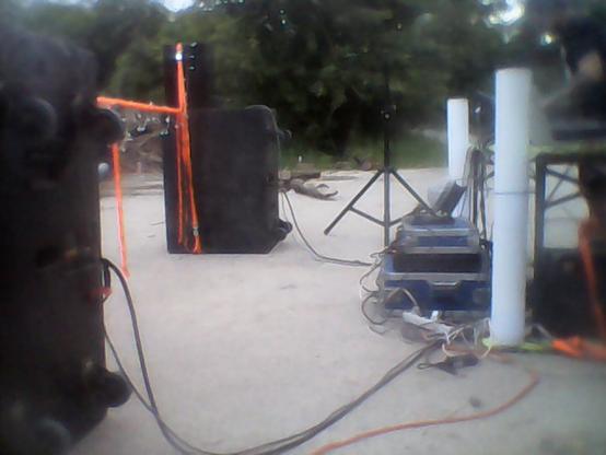You see the side view of a technical setup for a techno dance party. On the right side there is three cases stacked with controller and two amplifiers. Two thick loudspeaker cables are leading to the pictures left showing the loudspeaker stacks bundled by a orange strap and another to mimic a visible barrier infront of the DJ cockpit under a cheap party tent.