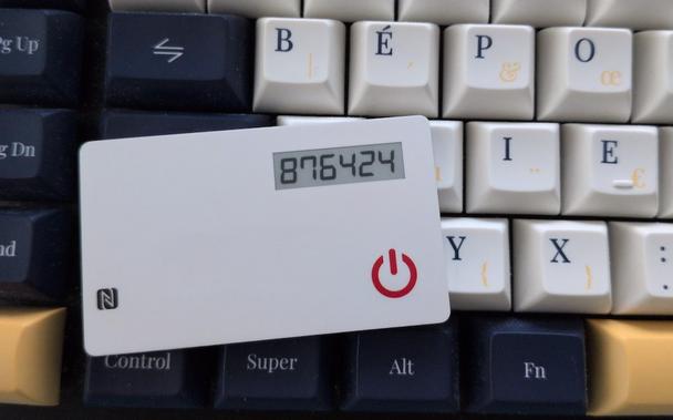 a Token2 miniOTP-3-i card laying on a keyboard for scale (and to brag about my BÉPO keyboard).
The card is way smaller than a credit card. It’s white and brand-less. On the left a tiny NFC logo is printed, the display is on the top right and the on/off button is on the botton right.