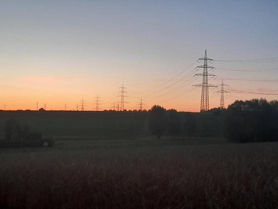 Eine leicht hügelige Landschaft mit Feldern, Wiesen, Bäumen und Hecken in der Dämmerung. Der Himmel ist vom Sonnenuntergang leuchtend Orange. Über die Landschaft ziehen sich mehrere Hochspannungstrassen und lokale Stromleitungen.