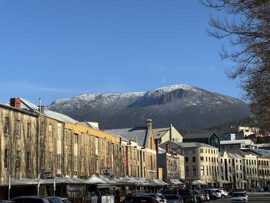 Another angle of the same view, this one cropping out the cars but revealing more of the snow capped Kunanyi and the ridgeline to its east. The shadows of leafless trees are on the buildings, various natural stone or pale yellow/cream. In colour.