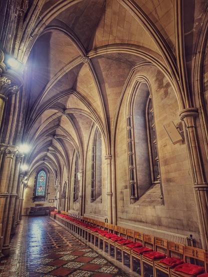 A long, high-angle interior view of the main corridor of Christ Church Cathedral in Dublin. The vaulted stone ceiling with ornate ribbing stretches down the length of the hall, which is illuminated by a soft, warm light from unseen sources. Tall, arched windows line the right side of the corridor, while a row of wooden chairs with deep red cushions extends along the right wall. The floor is tiled with a decorative pattern, reflecting the light from the left side of the image, where a stained-glass window can be partially seen. The overall mood is one of quiet solemnity and historical grandeur.