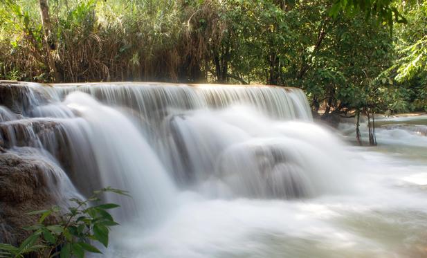 Photo shows a lower river fall with multiple streams and short falls The photo technic used slows down the water and gives the water flow a texture like textile fabric. It is placed within a jungle with trees and large trunks, and thick green foliage. Behind the heavy watery fog, it is a sunny day.