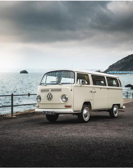 Das Bild zeigt einen klassischen, cremefarbenen Volkswagen Transporter, der an einem gepflasterten Weg am Meer geparkt ist. Der Transporter hat die charakteristischen runden Scheinwerfer und das Volkswagen-Logo auf der Front. Die Windschutzscheibe ist mit einem Muster verkleidet, und die Fenster sind mit Vorhängen ausgestattet. Der Fahrzeugkennzeichen lautet "KUM 687F". Im Hintergrund ist das Meer zu sehen, mit einem bewölkten Himmel und einer felsigen Küste im Hintergrund. Ein Stück weiter im Meer ist ein Felsen zu erkennen. Der Weg ist von einem Metallgeländer gesäumt, das zum Meer führt. Die Atmosphäre des Bildes ist ruhig und nostalgisch, mit einem Hauch von Abenteuer.