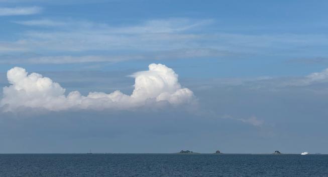 The North Sea under some clouds. On the horizon the buildings on Hallig Nordstrandischmoor are visible. A white Ferry is on the right side.