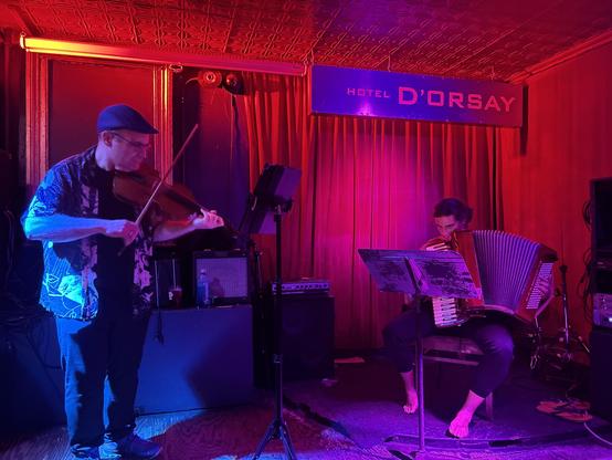 Ljova and Simone Baron performing at Barbes in Brooklyn. Ljova plays a fadolín (six string violin) and Simone plays an accordion. The background is dark red.