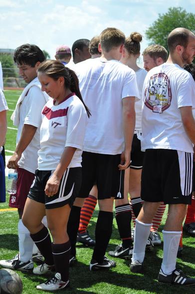 A group of people is standing on a soccer field, dressed in athletic clothing. Some individuals are wearing white shirts, while others have different colored shorts and socks. The scene captures a moment before or after a game, with a soccer ball visible.