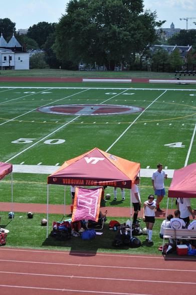 A sports field featuring a Virginia Tech tent and various soccer players. Soccer equipment and bags are visible near the tent, with a discernible field marking at the center. The background includes trees and a few buildings.